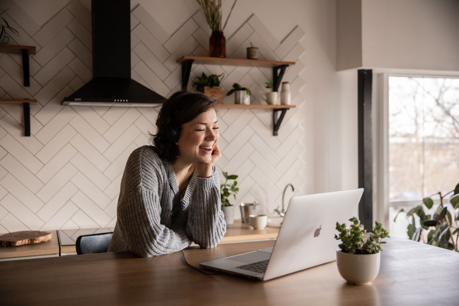 A working-age adult smiling on a laptop video call at a bright kitchen counter, reaching care from home